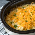 Close-up of hot, bubbling slow cooker crockpot cheesy potatoes casserole topped with melted golden cheddar cheese and green chives, served in a white dish.